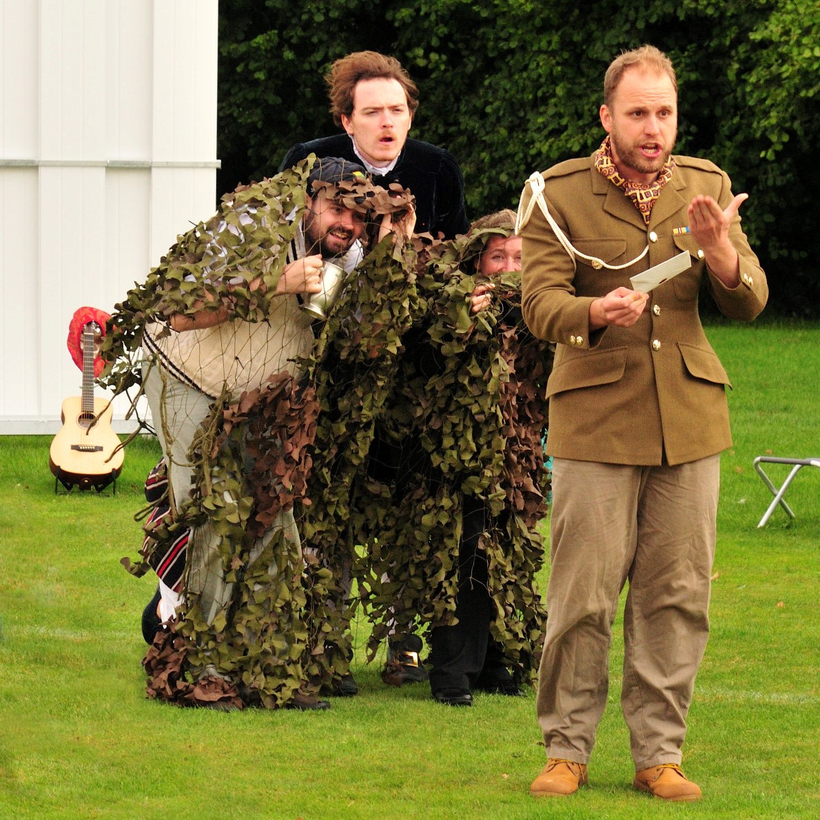 Outdoor photo from the 2021 production of Twelfth Night showing Sir Toby, Feste and Maria hiding in camoflage netting behind Malvolio who reads from his letter