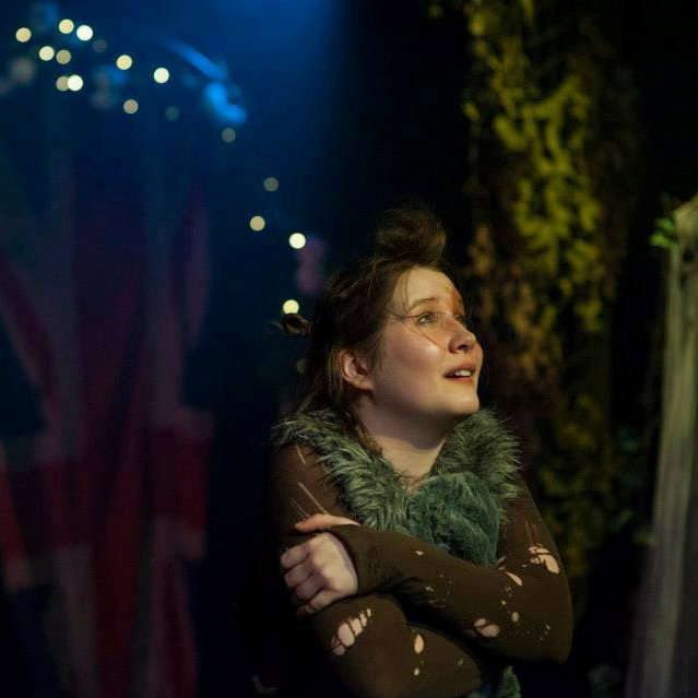 Puck hugging themselves arms crossed with forest and fairy lights in background